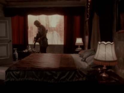 Chuck Wagner hat ein Gewehr in der Hand und steht im Schlafzimmer neben einem in rot bezogenen Luxusbett