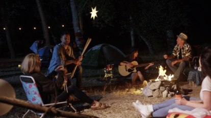 Camper sitzen um ein Lagerfeuer, eine von ihnen hat eine Gitarre